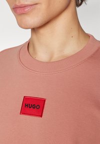 Gros plan sur une personne portant un sweat-shirt rose poussiéreux avec un patch carré rouge et "HUGO" en lettres noires sur la poitrine.
