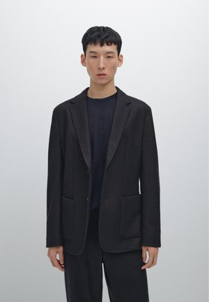Schwarzes maßgeschneidertes Blazer aus strukturiertem Stoff, mit Reverskragen, zwei vorderen Taschen und einem lockeren Schnitt über einem dunklen Hemd.