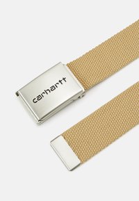 Curea împletită de culoare nisipie, cu o cataramă din metal argintiu pe care este gravat logo-ul "carhartt". Capetele au accente argintii contrastante. Material durabil și texturat.