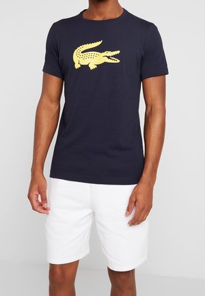 Homme portant un t-shirt bleu marine avec un grand motif jaune de crocodile et un short blanc, debout devant un fond clair.
