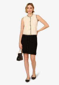 Krémový bezrukávový top s texturovanou látkou a zlatými knoflíčky, kombinovaný s upnutou černou tweedovou sukní s předními kapsami a třepícími se okraji.