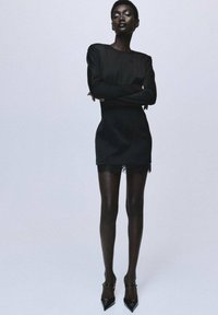 Vestido mini negro con mangas largas, escote transparente y ribete de encaje en el dobladillo, combinado con tacones negros de punta y medias opacas.