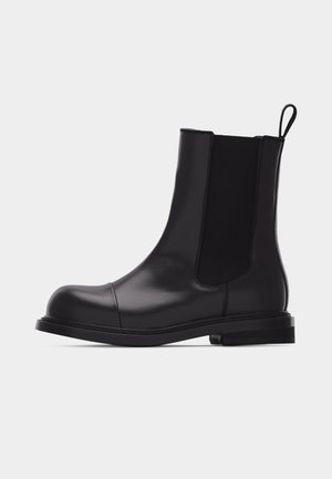 Fekete bőr bokacsizma elasztikus oldalpanelekkel, lekerekített orr-résszel, hátul húzófüllel és alacsony sarkú, oldalról fehér háttér előtt bemutatva.