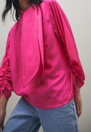 Person trägt eine leuchtend pinke Bluse mit langen Ärmeln, gerafften Ärmelabschlüssen und einer großen Schleife am Hals, kombiniert mit hellblauen Jeans.
