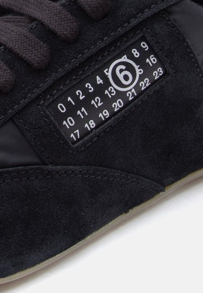 Baskets noires combinant suède et matériaux synthétiques, avec une surface texturée et une étiquette graphique affichant des chiffres et un cercle.