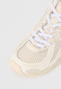 Chaussure de sport beige avec un extérieur en mesh, des lacets blancs et une semelle rembourrée. Présente des lignes de design subtiles et des accents texturés pour une meilleure adhérence.