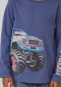 Niebieska koszula z długim rękawem z dużą grafiką kolorowego monster trucka z przesadnie dużymi oponami i szczegółowym widokiem z przodu.