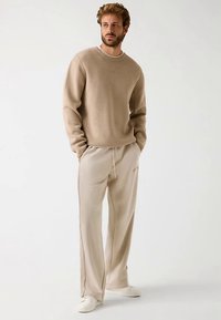 Beiger Pullover mit erhabenem "GUESS"-Logo, kombiniert mit hellbeigen Sweatpants mit Kordelzug und Ton-in-Ton-Branding, getragen mit weißen Sneakern.