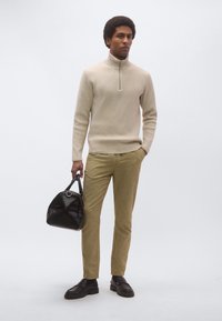 Μπεζ πλεκτό πουλόβερ με φερμουάρ, ανοιχτό καφέ υφασμάτινο παντελόνι, μαύρα δερμάτινα παπούτσια και μαύρη καπιτονέ τσάντα χειρός που κρατιέται στο δεξί χέρι.