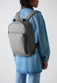 Mochila gris hecha de material suave, con un bolsillo frontal con cremallera, correas acolchadas y líneas limpias. Lleva puesta sobre una chaqueta de mezclilla y pantalones.