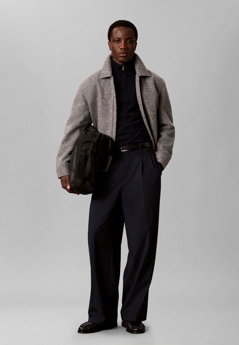 Homme portant une veste grise, un col roulé noir et un pantalon large noir tenant un sac noir, debout devant un fond gris uni.