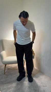 Hombre con cabello trenzado, vestido con una camiseta blanca y pantalones negros, de pie con las manos en los bolsillos sobre un suelo alfombrado, cerca de un sillón blanco y una mesa pequeña.