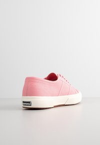 Niskie różowe trampki z płótna z białą gumową podeszwą, sznurowadłami oraz metką marki "Superga" na pięcie, przedstawione od tyłu na białej powierzchni.
