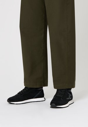 Sneakers in suede neri con suole bianche e lacci neri. Le scarpe presentano un accento laterale in contrasto. Abbinate a pantaloni larghi verde oliva.