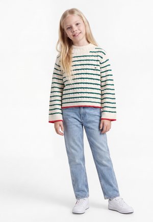 Ung pige med langt blondt hår iført en cremefarvet sweater med grønne striber, lyseblå jeans og hvide sneakers, der står på hvid baggrund.
