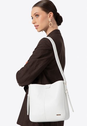 Femme portant un blazer sombre et un grand sac à épaule en cuir blanc, portant des boucles d'oreilles créoles dorées, les cheveux tirés en arrière en un chignon soigné sur fond blanc.