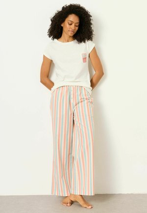 Frau mit lockigem Haar, die ein weißes kurzärmliges Hemd und pastellfarbene, gestreifte, weit geschnittene Hosen trägt, barfuß an einer weißen Wand stehend.