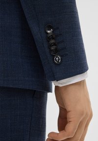 Maniche di giacca di un completo blu navy a trama, con quattro bottoni neri e dettagli di cucitura, e un polsino di camicia bianca che spunta da sotto.