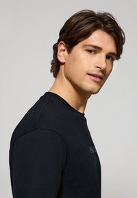 Svart t-shirt med kort ärm och rund halsringning, med subtila logodetaljer i en något ljusare nyans, slät tygtextur.