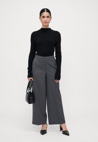 Vrouw in een zwart geribbeld sweater, grijs gestreepte wijde broek, zwarte hakken, met een zwarte handtas voor een witte achtergrond.