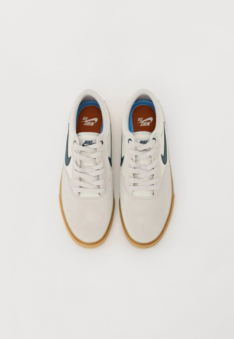 Zapatillas Nike de ante blanco, con un swoosh en azul marino, suela de goma y detalles naranjas en las plantillas. Diseño de cordones con forma de punta redondeada.