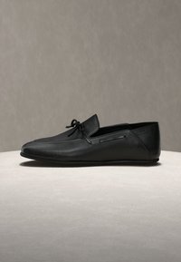Mocassin en cuir noir à enfiler avec un petit nœud sur le dessus, doté d'une semelle lisse et de détails de couture discrets.