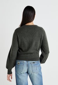 Temno zelena pletena majica z okroglim izrezom in napihnjenimi rokavi, v kombinaciji z svetlo modrimi jeans hlačami, ki imajo na hrbtni strani oznako blagovne znamke.