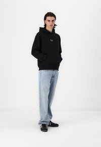 Svart hettegenser med frontlomme, kombinert med lyseblå baggyjeans. Modellen har på seg svarte joggesko med hvite detaljer, og står mot en hvit bakgrunn.
