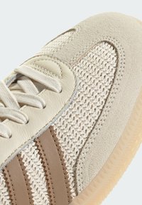 Sneaker beige clair avec des accents en textile tissé et en cuir ; comporte trois rayures marron, profil bas, et une semelle en caoutchouc texturée.
