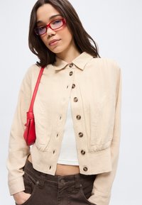 Νεαρή γυναίκα με κόκκινα γυαλιά, μπεζ μπουφάν με κουμπιά, λευκό τοπ, σκούρο παντελόνι και κόκκινη τσάντα στον ώμο, σε λευκό φόντο.