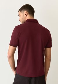 Vyno raudonos spalvos polo marškinėliai su tekstūruotu apykakliu ir trumpomis rankovėmis. Lygus audinys ir aptemptas siluetas, šiek tiek pailginta nugaros dalis.