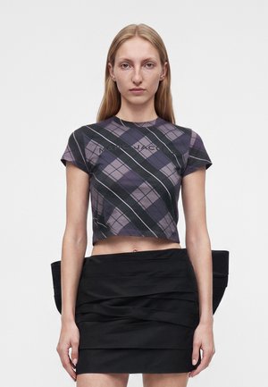 Jeune femme aux longs cheveux blonds portant un t-shirt court à carreaux violets et une mini-jupe noire froncée sur un fond uni.