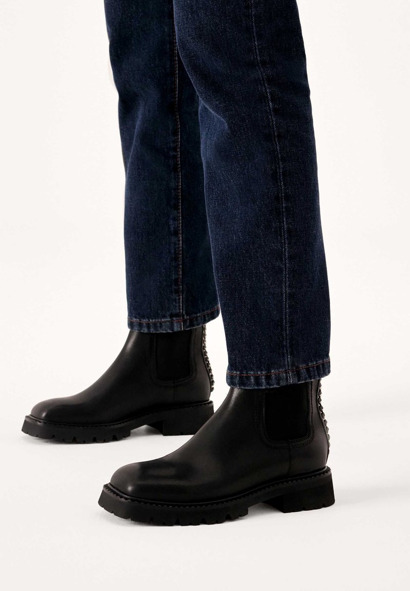 Bottes Chelsea en cuir noir avec semelles robustes portées avec un jean droit bleu foncé sur un fond clair uni.