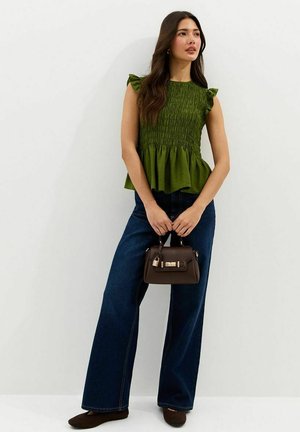 Vrouw draagt een groene mouwloze top met ruches, wijde donkerblauwe jeans, bruine platte schoenen, en houdt een kleine bruine handtas vast.