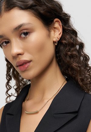 Goldene Halskette mit geschwungenem, strukturiertem Balkenanhänger. Modell hat lockiges Haar und trägt passende goldene Creolen. Schwarzes Oberteil mit Kragen.