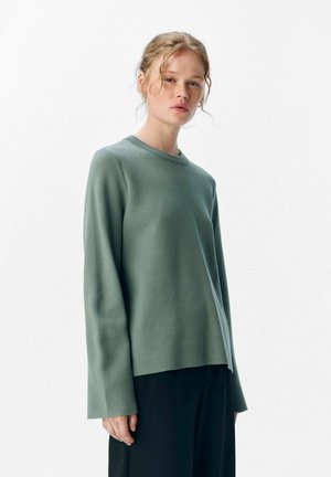 Vrouw met licht krullend haar draagt een losse groene trui met lange mouwen en zwarte broeken, staande tegen een eenvoudige witte achtergrond.