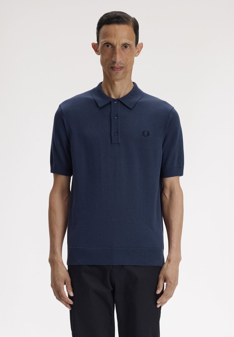 Homme debout portant un polo à manches courtes bleu marine et un pantalon noir sur un fond blanc uni.