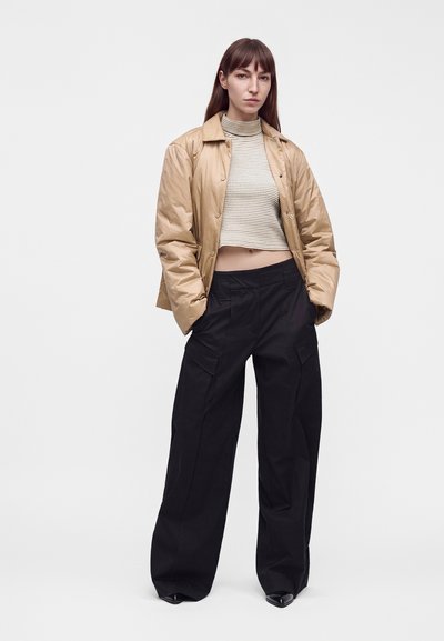 Femme debout, les mains dans les poches, portant une veste beige, un col roulé court rayé et un pantalon cargo noir à jambes larges sur fond blanc.