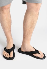 Schwarze Flip-Flops mit flacher Sohle, weichen Riemen und Zehensteg. Die Riemen haben eine strukturierte Oberfläche für besseren Halt.