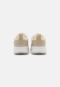 Beige sneakers with metallic gold accents, a textured tan back, white sole, and branding on the heel. Smooth upper and cushioned collar.