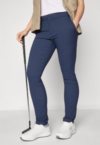 Navy golf pants made of stretchable fabric with a slim fit, featuring pockets and paired with white sporty shoes and a golf club.
