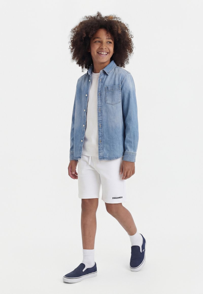Enfant souriant marchant, portant une chemise en denim bleu clair sur un t-shirt blanc, un short blanc, des chaussettes blanches et des chaussures bateau bleu marine.