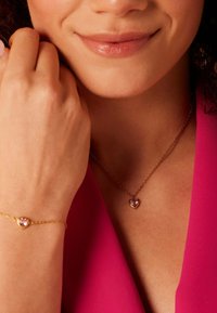 Gouden sieraden omvatten een hartvormige hangerketting en armband, met roze edelstenen op een delicate ketting tegen een roze achtergrond.