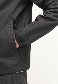 Personne portant une veste noire avec des poignets élastiques, main dans une poche latérale, et un pantalon sombre à motifs visible sous la veste.