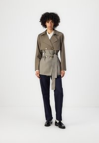 Dobbeltradet trenchcoat i olivengrønn og grå stoff, med belte i midjen, store slag og knapper i gulltone. Kombinert med marineblå bukser.