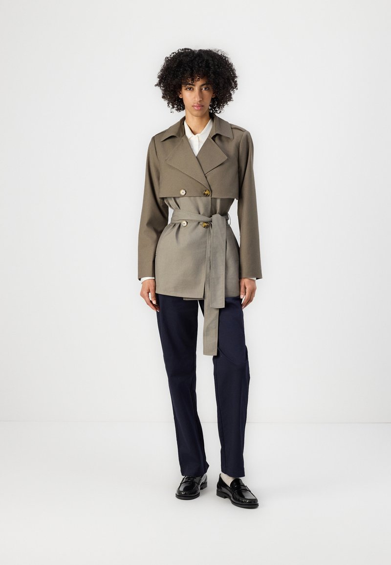 Dobbeltradet trenchcoat i olivengrønn og grå stoff, med belte i midjen, store slag og knapper i gulltone. Kombinert med marineblå bukser.