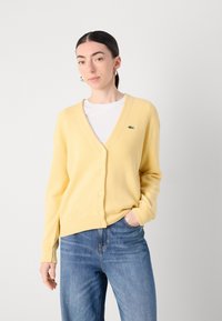 Žlutý cardigan vyrobený z texturované látky, s výstřihem do V, knoflíkovým zapínáním a malým zeleným logem. V kombinaci s modrými džínami.