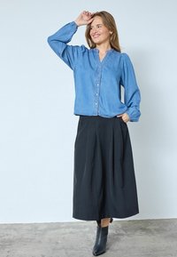 Camicia in denim chiaro con bottoni, dalla vestibilità rilassata, maniche lunghe e scollatura a V, abbinata a una gonna nera fluida plissettata e stivaletti.