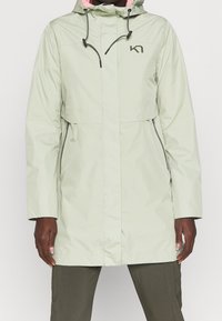 Ljusgrön vattentät jacka med huva, dragkedjefickor och rosa inredning. Har en logotyp på bröstet. Långa ärmar.