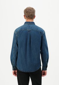 Un homme aux cheveux courts portant une chemise en denim bleu à manches longues et un pantalon noir, de dos, devant un fond blanc uni.
